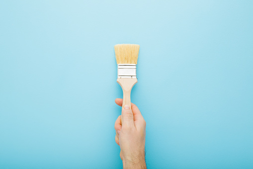 Young adult man hand holding new paint brush on light blue table background. Pastel color. Closeup. Preparation for repair work of home. Top down view.