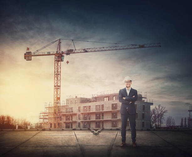 istock Man contractor or architect wearing protective helmet stands confident on the rooftop in front of a construction site with working tower crane. Engineer with hardhat inspecting the safety at work 1372386671