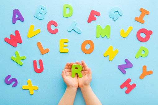 Toddler boy hands holding R letter. Colorful letters on light blue table background. Pastel color. Time to learning English alphabet. Point of view shot. Closeup. Top down view.