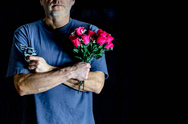 Man holding flowers and gun with arms crossed, wearing blue t-shirt, on black background.The man has mixed feelings
