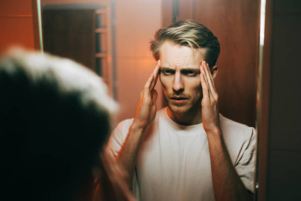 istock Young exhausted caucasian man standing in front the mirror and looking on himself, and touching the temples. 1372400337