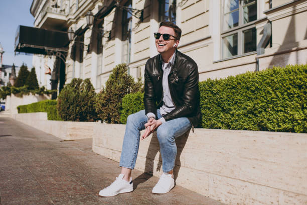 Full length young happy amazed fun smiling caucasian happy man 20s wear black leather jacket eyeglasses sitting near bulding and bushes resting in city downtown outdoors Concept of urban lifestyle. joven de cuerpo entero feliz asombrado diversión sonriente caucásico hombre feliz 20s usa anteojos de chaqueta de cuero negro sentado cerca de bulding y arbustos descansando en el centro de la ciudad al aire libre concepto de estilo de vida urbano