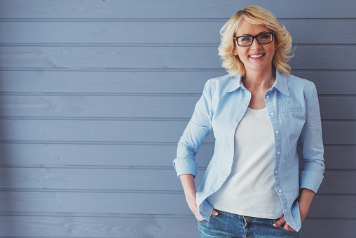 Beautiful mature woman in casual clothes and eyeglasses is looking at camera and smiling, standing against gray background