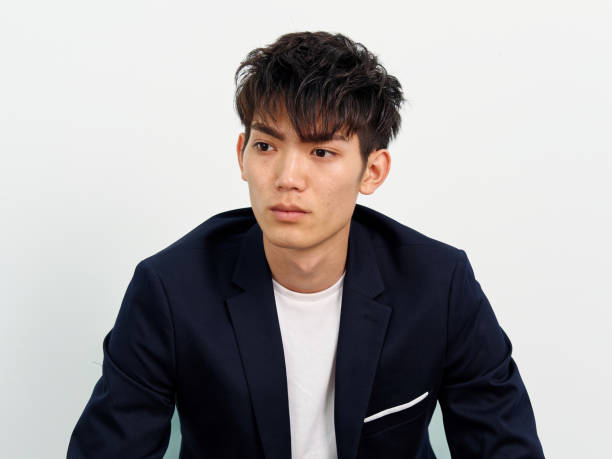 portrait of handsome chinese young man in dark blue leisure suit sitting in armchair and posing against white wall background. looking away seriously, front view. - human face chinese ethnicity close up men imagens e fotografias de stock