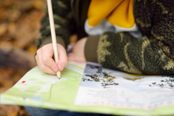 Little boy scout is orienteering in forest. Child is looking on map on background of teepee hut. Adventure, scouting and hiking tourism for kids. Little boy scout is orienteering in forest. Child is looking on map on background of teepee hut. Concepts of adventure, scouting and hiking tourism for kids. orienteering stock pictures, royalty-free photos & images