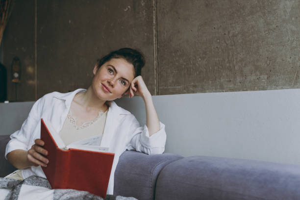 Young dreamful thoughtful minded woman in white clothes plaid sitting on soft grey sofa indoors apartment read novel book textbook prop up forehead Rest on weekend leisure quarantine stay home concept joven mujer soñadora de mente reflexiva con ropa blanca a cuadros sentada en un sofá gris suave apartamento interior leer libro de texto novedoso libro de texto apuntalar la frente descansar en el fin de semana ocio cuarentena quédate en casa concepto