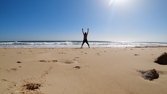 Man jumps for joy to finally go to beach on sunny afternoon with waves and golden sand