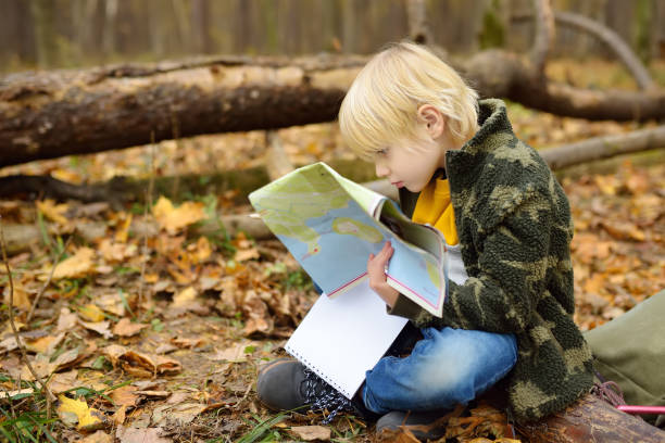istock Little boy scout is orienteering in forest. Child is sitting on fallen tree and looking on map on background of teepee hut. Adventure, scouting and hiking tourism for kids. 1372424014
