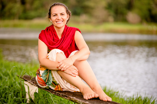 Portrait of a young woman smiling while sitting on a bench outside by a tranquil lake
