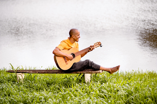 Smiling young man sitting on a bench by a lake and playing the guitar during a relaxing day in summer