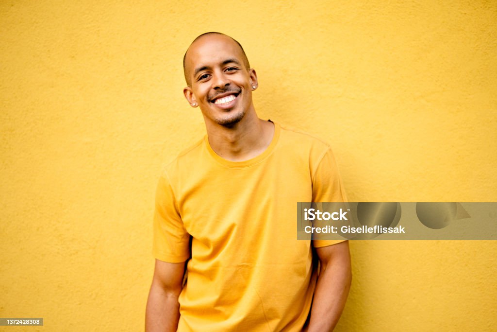 Young man smiling while wearing a yellow shirt by a yellow wall Portrait of a smiling young man wearing a yellow t-shirt leaning against a yellow wall outdoors Yellow Stock Photo