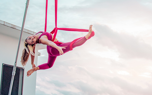 Beautiful healthy strong woman wearing sport bra, relaxing, exercise yoga fly in air, aerial hoop on outdoor rooftop in morning. There are background of sky, sunlight flare, copy space advertisement