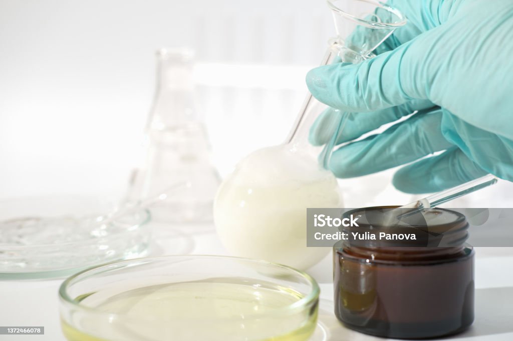 hand in rubber medical glove holding laboratory glassware with fermented cosmetic product, face cream with enzymes developed in a lab. hand in rubber medical glove holding laboratory glassware with fermented cosmetic product, face cream with enzymes developed in a lab Fermenting Stock Photo