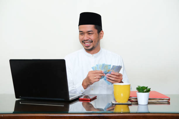 muslimischer asiatischer mann lächelt, während er sein geld vor dem laptop zählt - sharia stock-fotos und bilder