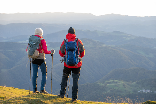 Rear view of couple standing with hiking poles on top of mountain and looking at view.