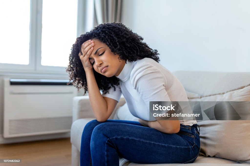 African American Woman in painful expression holding hands against belly suffering menstrual period pain, lying sad on home bed, having tummy cramp in female health concept Stomachache Stock Photo