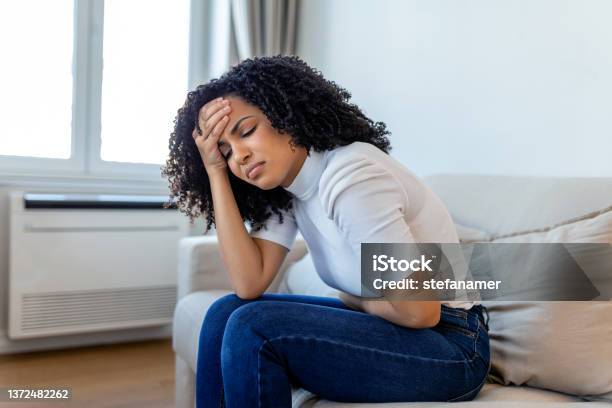 African American Woman In Painful Expression Holding Hands Against Belly Suffering Menstrual Period Pain Lying Sad On Home Bed Having Tummy Cramp In Female Health Concept Stock Photo - Download Image Now