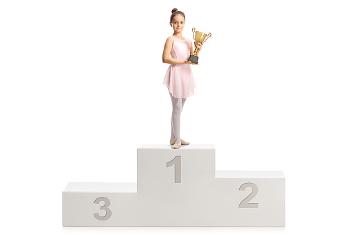Ballerina in a pink dress holding a gold trophy cup on a winner podium isolated on white background