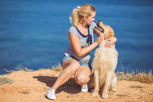 A girl trains a retriever dog in the summer on a deserted beach by the sea.Cheerful pretty young woman hugs her dog on the beach.  Labrador retriever and girl playing together on coastline. Lifestyle moment. Friendship between people and animals.