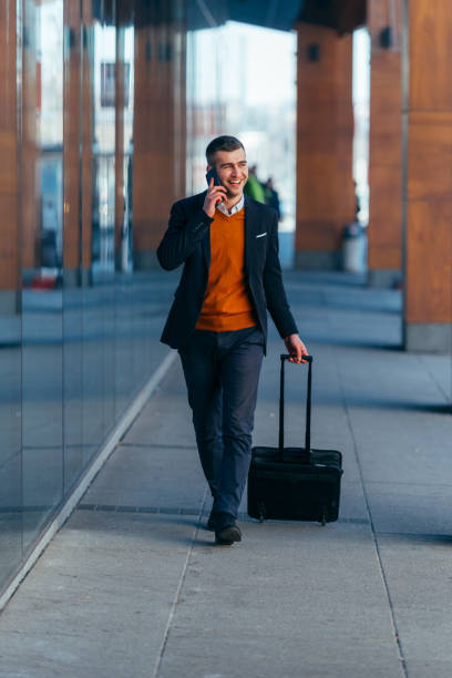 istock Young businessman walking through a station while talking on the phone and pulling his travel bag. 1372502039