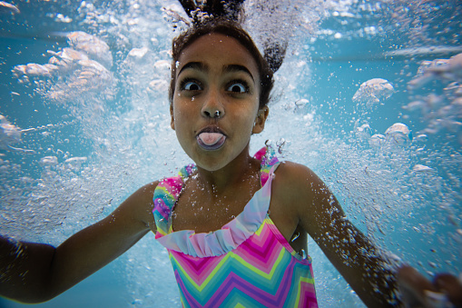 A young girl wearing swimwear, swimming underwater in a swimming pool. She is looking at the camera and sticking her tongue out. There are bubbles surrounding her.