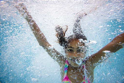 Close-up of a young girl wearing swimwear, swimming underwater in a swimming pool while looking at the camera and smiling with her arms raised. She is surrounded by bubbles.