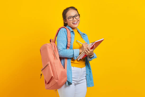 istock Smiling young Asian woman student in denim clothes with backpack, writing notes in notebook isolated on yellow background. Education in high school university college concept 1372505225