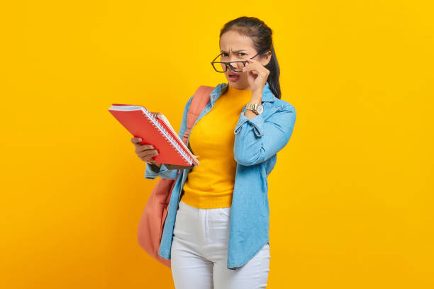 istock Pensive beautiful young Asian woman student in denim clothes with backpack holding notebook and looking at camera isolated on yellow background. Education in high school university college concept 1372505243