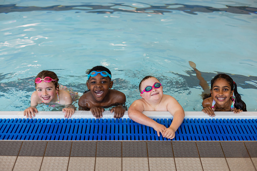 A group of children leaning and gripping onto poolside while playing in the swimming pool in Boldon, North East England. They are splashing smiling while looking at the camera. Three of them are wearing swimming goggles.