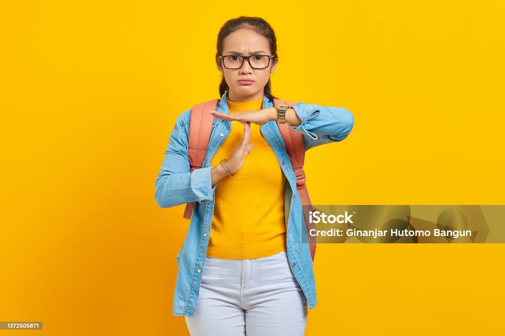 Beautiful young Asian woman student in denim outfit with backpack showing time out gesture with hands isolated on yellow background. Education in university college concept 20-29 Years Stock Photo