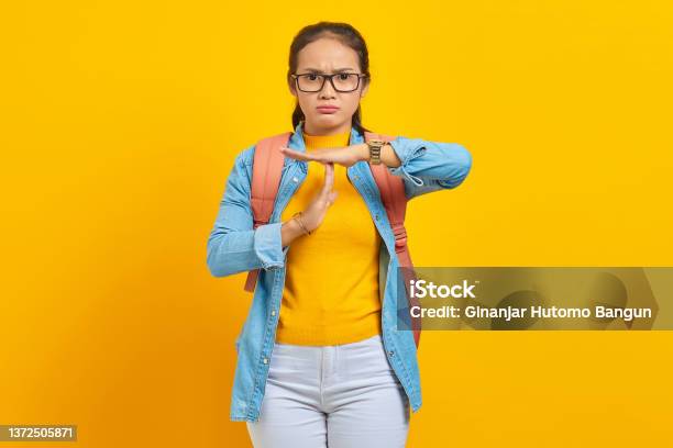 Beautiful Young Asian Woman Student In Denim Outfit With Backpack Showing Time Out Gesture With Hands Isolated On Yellow Background Education In University College Concept Stock Photo - Download Image Now