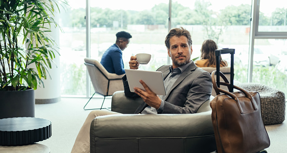 Smiling business man reading something on a tablet and holding cup of coffee while sitting in armchair and waiting for flight at airport terminal longe.