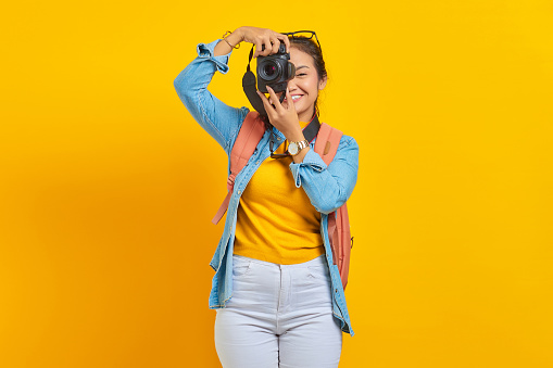 Portrait of cheerful young Asian woman with backpack taking photo at camera profesional isolated on yellow background