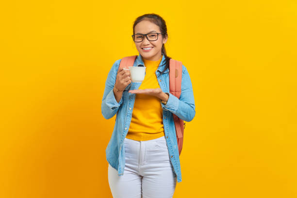 istock Excited beautiful young Asian woman student in denim clothes with backpack while showing cup of coffee with palms isolated on yellow background 1372507881