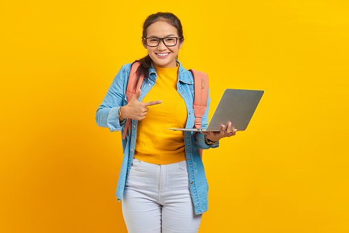 Portrait of smiling young Asian woman student in casual wear with backpack pointing at laptop with finger isolated on yellow background. Education in college university concept