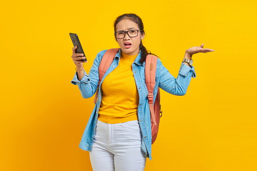 Portrait of  confused young Asian woman student in casual clothes with backpack holding mobile phone and saying no with palms isolated on yellow background.  Education in college university concept