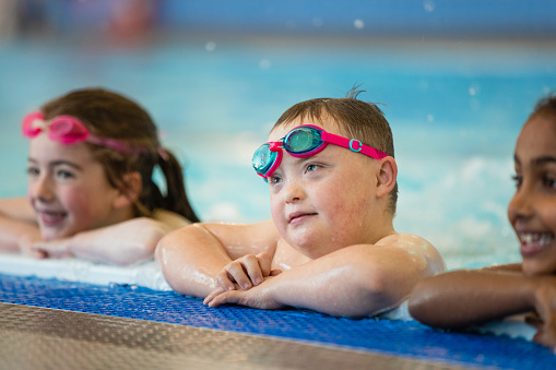 Two female children and a boy leaning against the poolside while playing in the swimming pool in Boldon, North East England. They are looking in front of them and smiling. The main focus is the boy wearing goggles and looking at something out of shot.