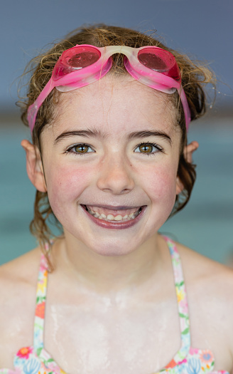 A young girl wearing swimming goggles, looking at the camera and smiling at a swimming pool in Boldon, North East England. She is wet after having a swim in the pool.