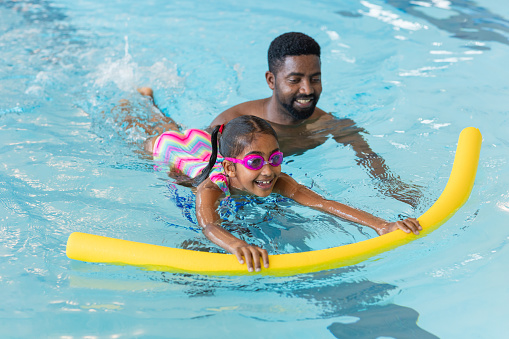 A man teaching his young daughter how to swim in a swimming pool in Boldon, North East England. The daughter is wearing goggles and is using a woggle to help support her in the water.
