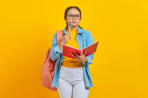Beautiful young Asian woman student in casual clothes with backpack holding book and looks serious thinking about  creative idea isolated on yellow background