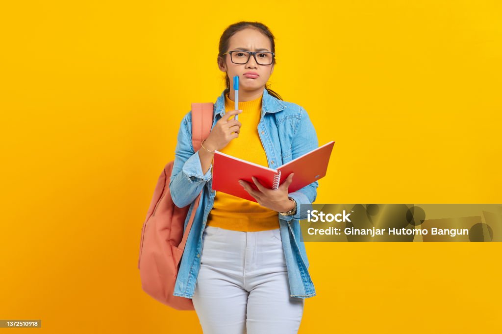 Beautiful young Asian woman student in casual clothes with backpack holding book and looks serious thinking about  creative idea isolated on yellow background Learning Stock Photo