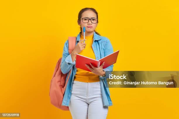 Beautiful Young Asian Woman Student In Casual Clothes With Backpack Holding Book And Looks Serious Thinking About Creative Idea Isolated On Yellow Background Stock Photo - Download Image Now