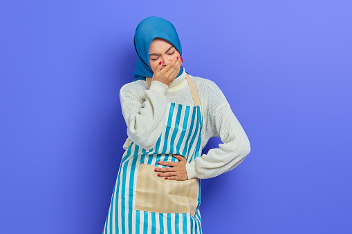 Portrait of sick young housewife woman in hijab and stiped apron,  having abdominal pain, put your hand on your stomach, feel nauseous and want to throw up isolated on purple background
