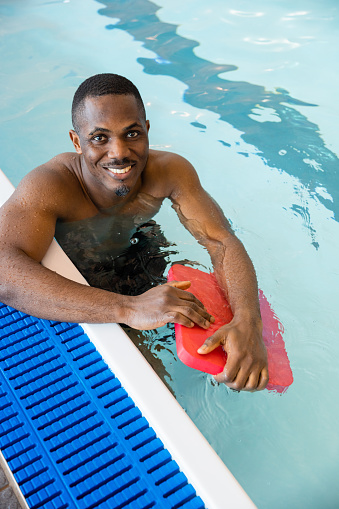 A mid adult man in a swimming pool, leaning on the poolside, holding a pool float while looking up at the camera and smiling. He is in Boldon, North East England.