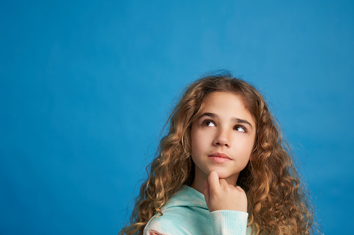 Portrait of dreamy and curious looking girl holding hand on chin isolated on blue background.