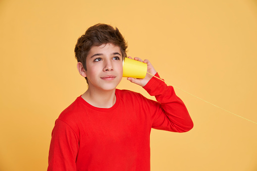 a boy listening through a paper cup tied to a thread isolated on yellow background.