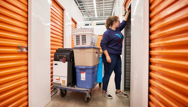 Woman storing her belongings in self storage unit Woman opening the shutter of a self storage unit with a trolley full of goods and items in the aisle of self-storage building storage compartment stock pictures, royalty-free photos & images