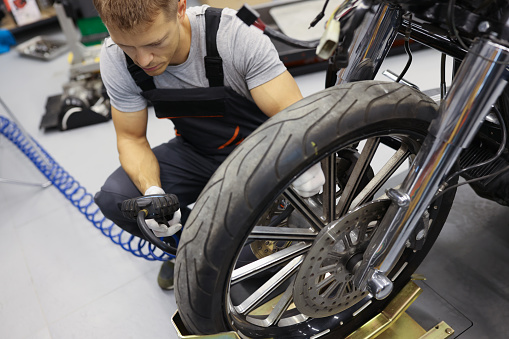 In the garage, a man in overalls sits near the wheel of a motorcycle, close-up. Checking the air pressure in the wheel tire