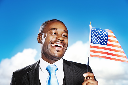 This smiling African American man could be a patriotic businessman or perhaps a politician as he enthusiastically waves the American national flag against a summer sky with puffy clouds and copy space,