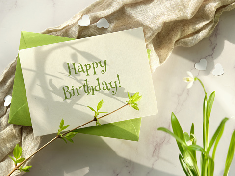 Happy Birthday card Spring background. Paper card and green envelope. Fresh leaves, snowdrop flowers. White paper heart confetti on off white linen and marble stone background.